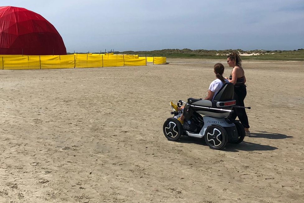 Een foto van Caroline, een blije vrouw van middelbare leeftijd, rijdend op een scootmobiel. Ze glimlacht breed terwijl ze haar scootmobiel langs een pittoresk parkpad stuurt.